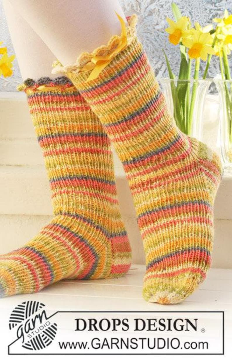 May include: A pair of mid-calf knit socks featuring a multi-colored striped pattern in shades of yellow, orange, pink, and blue. The socks have a scalloped crocheted top edge accented with a thin yellow ribbon tied in a small bow at the front.