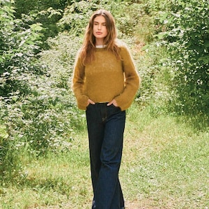 May include: A woman in a mustard-coloured jumper and dark blue wide-leg jeans stands outdoors. The jumper has a crew neck and long sleeves. The jeans are a classic denim style. The background features lush greenery and sunlight, creating a natural setting.