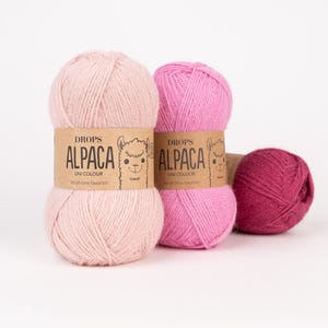 May include: Three skeins of yarn in shades of light pink, medium pink, and dark magenta. Each skein features a tan paper label with the text DROPS ALPACA UNI COLOUR and a small illustration of an alpaca face. The yarn has a soft, fibrous texture.