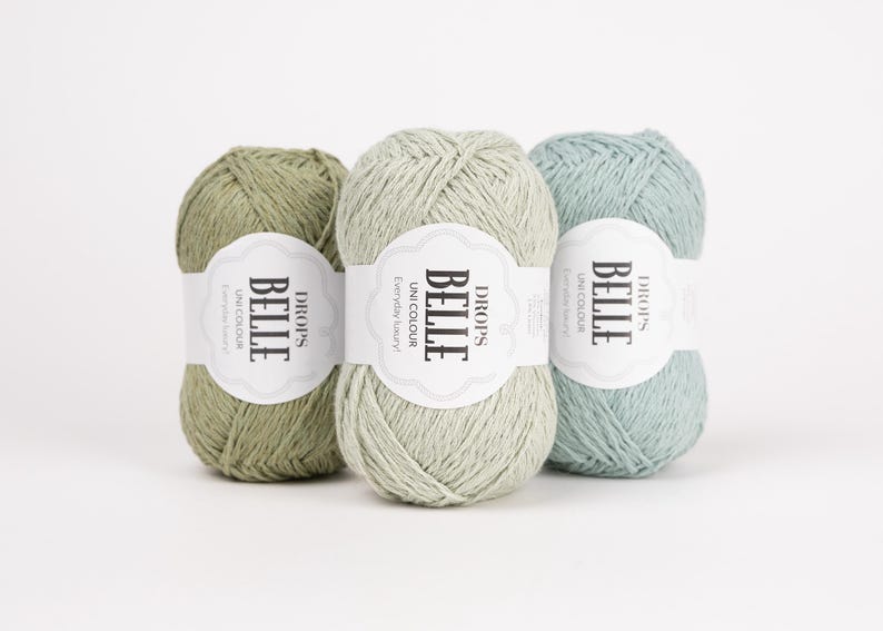 May include: Three skeins of Drops Belle yarn in shades of green and blue. Each skein has a white label with the text "BELLE" and "Unicolour Everyday luxury!" The yarn is arranged against a white background.