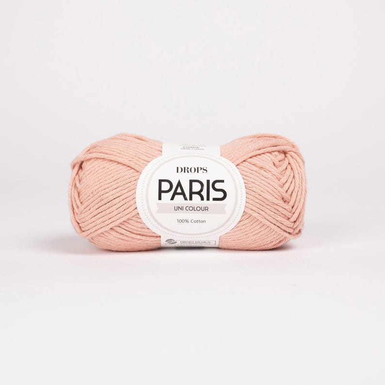 K&ouml;nnte beinhalten: Ein Kn&auml;uel Drops Paris Uni Colour Garn in einem zarten, hellrosa Farbton. Das Garn besteht aus 100 % Baumwolle und hat ein Etikett mit Markennamen und Produktinformationen. Geeignet f&uuml;r Strick- und H&auml;kelprojekte.