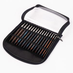 Puede incluir: Un estuche negro con cremallera y tapa transparente, que contiene un juego de agujas de tejer de madera. Las agujas son de varios tonos de marr&oacute;n y azul, con puntas de metal plateado. El estuche est&aacute; abierto, mostrando las agujas.