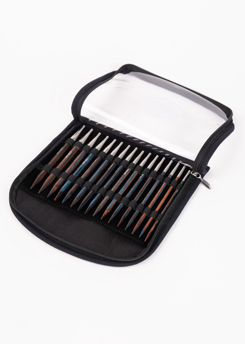 Puede incluir: Un estuche negro con cremallera y tapa transparente, que contiene un juego de agujas de tejer de madera. Las agujas son de varios tonos de marr&oacute;n y azul, con puntas de metal plateado. El estuche est&aacute; abierto, mostrando las agujas.