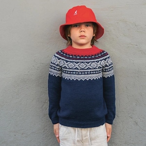 Puede incluir: Un niño con un sombrero de pescador rojo y un suéter azul marino con un patrón geométrico en blanco y rojo. El suéter tiene cuello y puños rojos. El niño también lleva pantalones cortos beige.