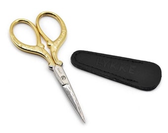 May include: Gold-colored embroidery scissors with a black leather case. The case has the word "LYKKE" embossed on it.