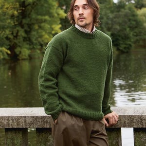 Könnte beinhalten: Ein grüner Strickpullover mit Rundhalsausschnitt. Der Pullover ist einfarbig und hat eine strukturierte Optik. Die Person trägt eine braune Hose. Der Hintergrund umfasst ein Gewässer und Bäume.