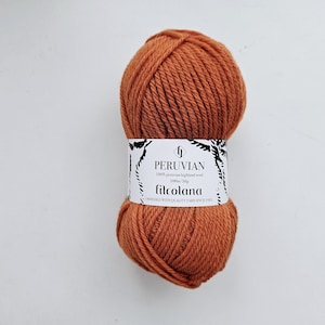 Op de afbeelding: Een bol roestoranje garen met een wit papieren label met de tekst PERUVIAN, 100% peruvian highland wool, 100m/50g, filcolana en OBSESSED WITH QUALITY YARN SINCE 1972. Het garen heeft een dikke, getwiste textuur en een effen kleur.