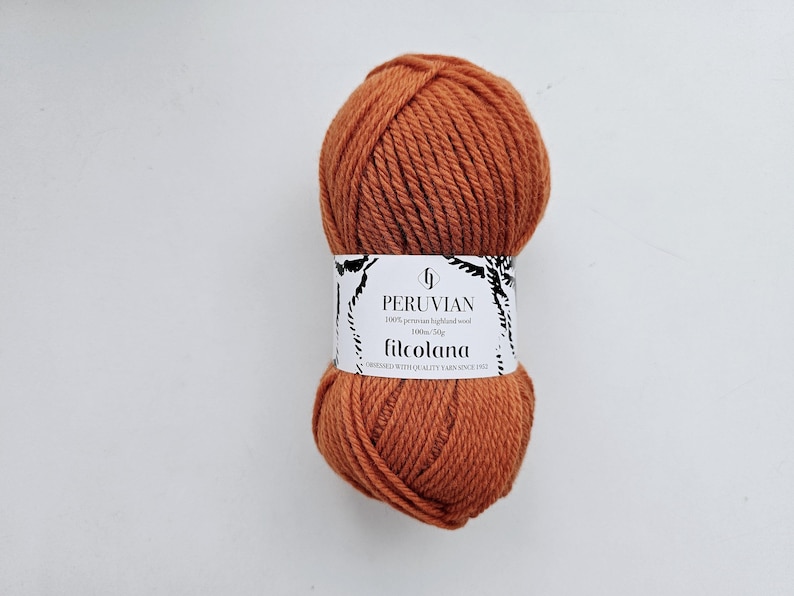 Op de afbeelding: Een bol roestoranje garen met een wit papieren label met de tekst PERUVIAN, 100% peruvian highland wool, 100m/50g, filcolana en OBSESSED WITH QUALITY YARN SINCE 1972. Het garen heeft een dikke, getwiste textuur en een effen kleur.