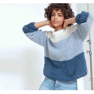 May include: A chunky knit sweater in shades of blue and white. The sweater features a color-block design with a dark blue bottom, a light blue middle, and a white top. The sleeves are a darker blue. The sweater is made of a thick, textured yarn.
