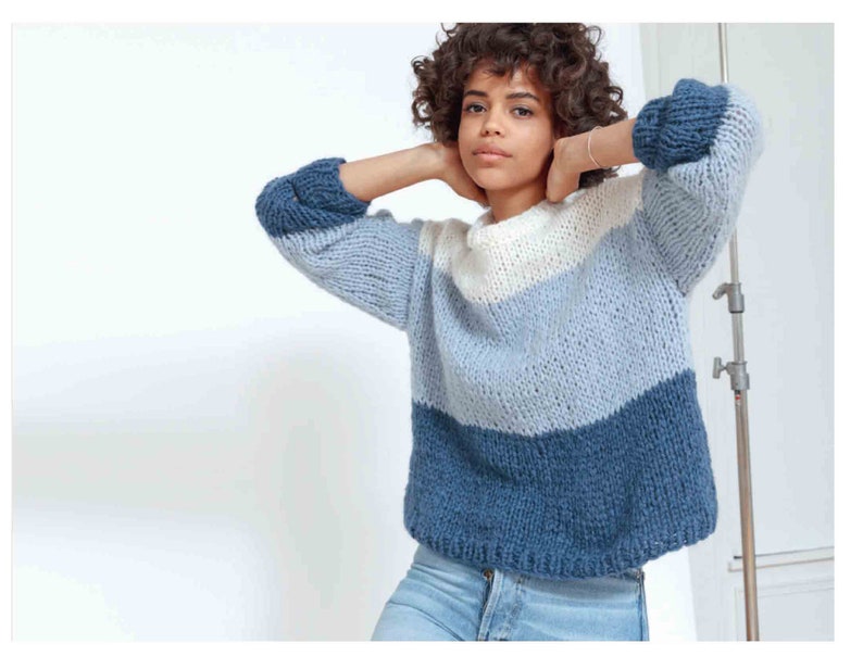 May include: A chunky knit sweater in shades of blue and white. The sweater features a color-block design with a dark blue bottom, a light blue middle, and a white top. The sleeves are a darker blue. The sweater is made of a thick, textured yarn.