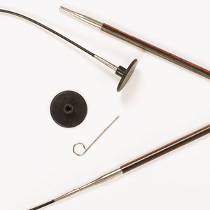 Puede incluir: Agujas de tejer y accesorios sobre un fondo blanco. El conjunto incluye dos agujas de tejer de madera con puntas met&aacute;licas, un cable negro, un tope negro y una peque&ntilde;a herramienta met&aacute;lica. Las agujas tienen un acabado marr&oacute;n oscuro.