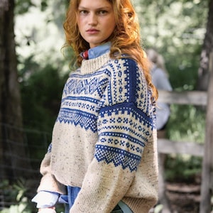 Puede incluir: Un suéter beige y azul estampado. El suéter tiene cuello alto y mangas largas con un diseño geométrico en azul. La modelo lleva debajo una camisa azul claro y vaqueros azules. El fondo es una escena borrosa al aire libre.