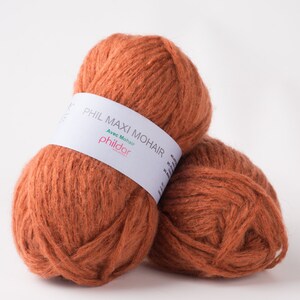 May include: Two balls of orange-brown yarn, labeled "PHIL MAXI MOHAIR" with "Avec Mohair" and the brand name "phildar" on a white label. The yarn has a fluffy texture, suitable for knitting or crochet projects.
