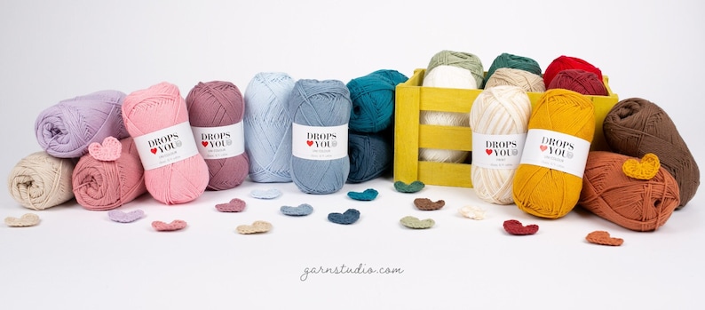 Puede incluir: Una colecci&oacute;n de madejas de hilo de varios colores, incluyendo rosa, azul y amarillo, dispuestas sobre una superficie blanca. El hilo est&aacute; etiquetado con "DROPS YOU" y acompa&ntilde;ado de peque&ntilde;os adornos en forma de coraz&oacute;n. Una caja de madera amarilla contiene hilo adicional.