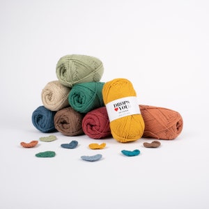 Puede incluir: Una colecci&oacute;n de ovillos de hilo coloridos en tonos verdes, beige, azul, marr&oacute;n, rojo y amarillo. El hilo est&aacute; dispuesto en una pila, con una etiqueta en un ovillo que dice "DROPS YOU". Peque&ntilde;os corazones de ganchillo est&aacute;n dispersos delante.