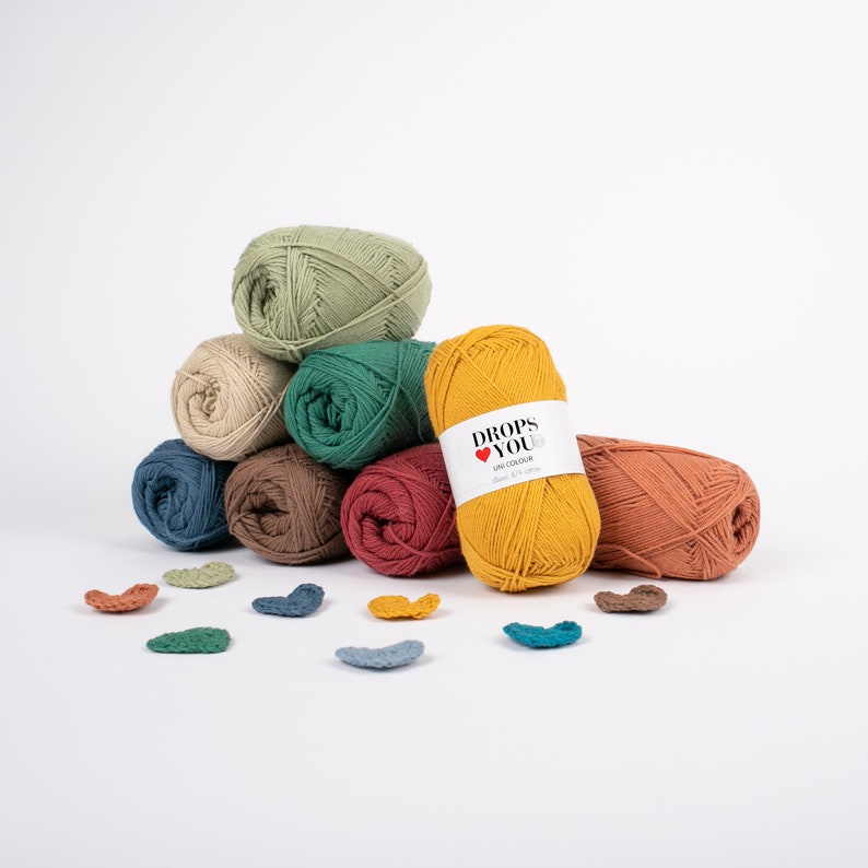 Puede incluir: Una colecci&oacute;n de ovillos de hilo coloridos en tonos verdes, beige, azul, marr&oacute;n, rojo y amarillo. El hilo est&aacute; dispuesto en una pila, con una etiqueta en un ovillo que dice "DROPS YOU". Peque&ntilde;os corazones de ganchillo est&aacute;n dispersos delante.