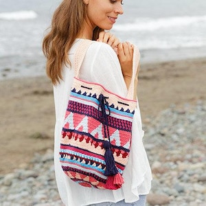 K&ouml;nnte beinhalten: Eine farbenfrohe, handgefertigte H&auml;keltasche mit einem geometrischen Muster in Rosa, Blau und Creme. Die Tasche hat einen Kordelzugverschluss und eine marineblaue Quaste. Die Tasche wird &uuml;ber der Schulter getragen.