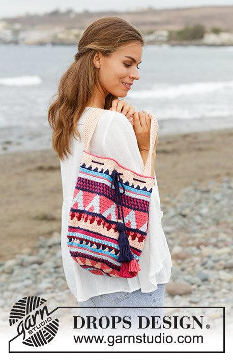 K&ouml;nnte beinhalten: Eine farbenfrohe, handgefertigte H&auml;keltasche mit einem geometrischen Muster in Rosa, Blau und Creme. Die Tasche hat einen Kordelzugverschluss und eine marineblaue Quaste. Die Tasche wird &uuml;ber der Schulter getragen.