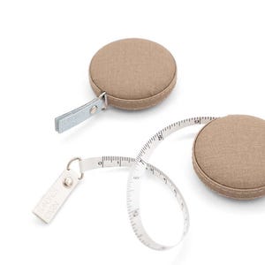 Puede incluir: Una cinta métrica de tela marrón con una cinta blanca y un cierre metálico. La cinta métrica está enrollada y el cierre está unido a una pequeña etiqueta rectangular que dice "SADDLES & GRAY".