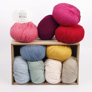 Puede incluir: Una colecci&oacute;n de diez ovillos de lana redondos en varios colores, incluyendo rosa, morado oscuro, rojo, azul, amarillo, azul claro, verde salvia, crema y beige, dispuestos dentro de una caja de madera con una etiqueta que dice DROPS COTTON MERINO UNI COLOUR.