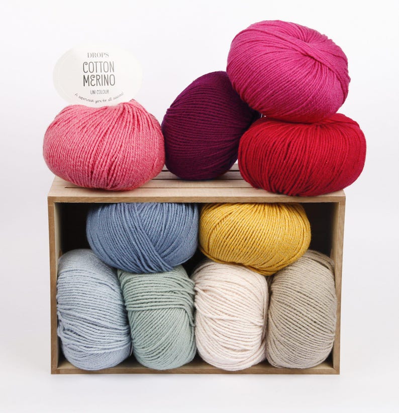 Puede incluir: Una colecci&oacute;n de diez ovillos de lana redondos en varios colores, incluyendo rosa, morado oscuro, rojo, azul, amarillo, azul claro, verde salvia, crema y beige, dispuestos dentro de una caja de madera con una etiqueta que dice DROPS COTTON MERINO UNI COLOUR.