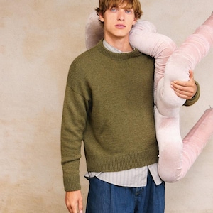 May include: An olive green crew neck sweater and blue denim trousers. The sweater is worn over a striped shirt. A large, pink, plush, worm-like object is held in the arms.