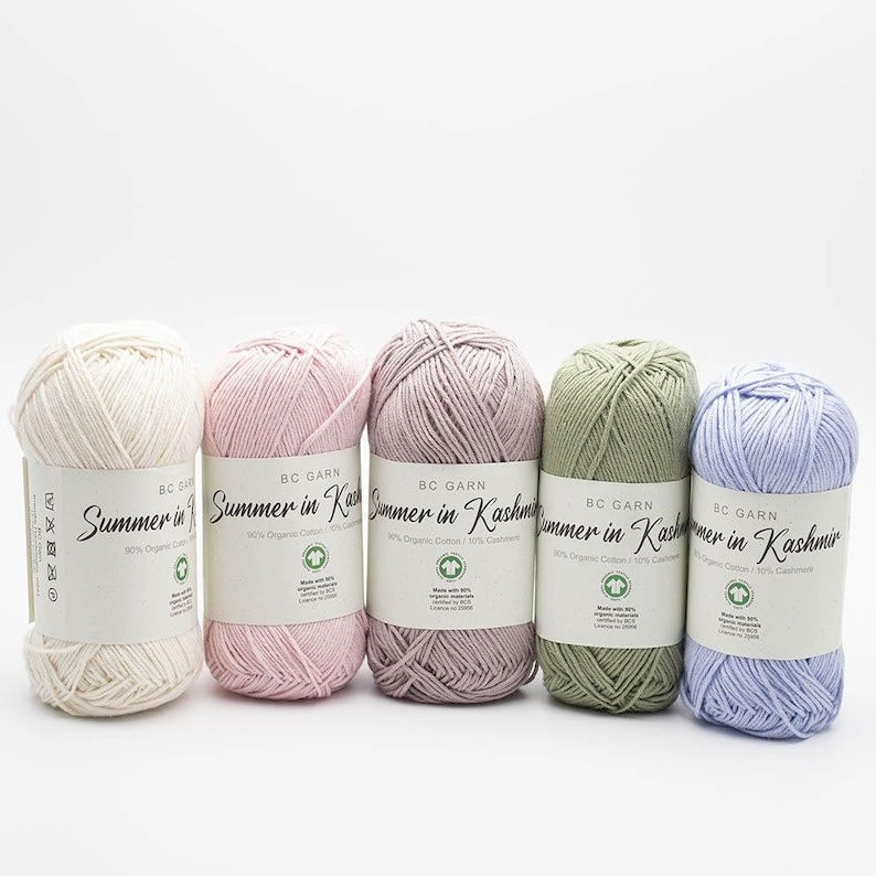Puede incluir: Cinco ovillos de lana en diferentes colores: blanco, rosa, morado claro, verde y azul claro. Cada ovillo tiene una etiqueta que dice "BC Garn Summer in Kashmir 90% algod&oacute;n org&aacute;nico / 10% cachemira".