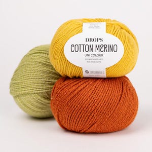Puede incluir: Tres ovillos de lana Drops Cotton Merino en amarillo, verde salvia y naranja quemado. El ovillo amarillo tiene una etiqueta de papel blanco con el texto "DROPS COTTON MERINO UNI COLOUR A superwash yarn for all seasons" y detalles.