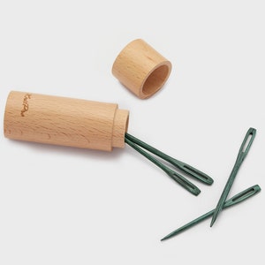 Puede incluir: Un juego de cuatro agujas de madera verde en una caja de madera. La caja es cilíndrica y tiene una tapa. Las agujas son afiladas y puntiagudas. La caja está etiquetada con "KnitPro".