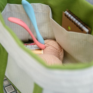 Puede incluir: Un bolso tote verde y blanco lleno de materiales de manualidades. En su interior hay dos ganchillos, uno rosa y otro azul, junto con una madeja de hilo y un peque&ntilde;o cuaderno. El bolso tiene el interior verde y el exterior blanco.