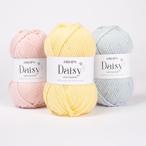 May include: Three skeins of yarn in pastel pink, pale yellow, and light blue. Each skein features a white paper label with the text DROPS Daisy UNI COLOUR 100% extra fine merino wool. The yarn is wound into a rounded, textured ball shape.