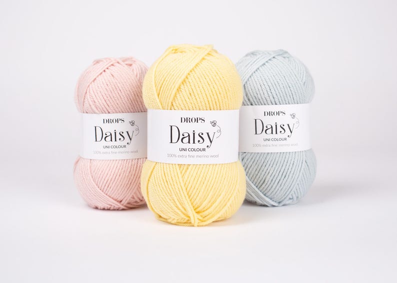 May include: Three skeins of yarn in pastel pink, pale yellow, and light blue. Each skein features a white paper label with the text DROPS Daisy UNI COLOUR 100% extra fine merino wool. The yarn is wound into a rounded, textured ball shape.