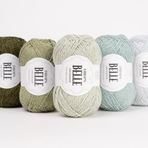 May include: Five skeins of Drops Belle yarn in shades of green and blue. Each skein has a white label with the text "BELLE" and "UNICOLOUR". The yarn is arranged in a row, showcasing the different colors available for knitting and crochet projects.