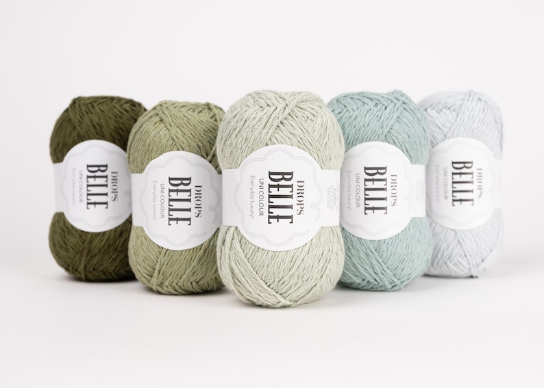 May include: Five skeins of Drops Belle yarn in shades of green and blue. Each skein has a white label with the text "BELLE" and "UNICOLOUR". The yarn is arranged in a row, showcasing the different colors available for knitting and crochet projects.