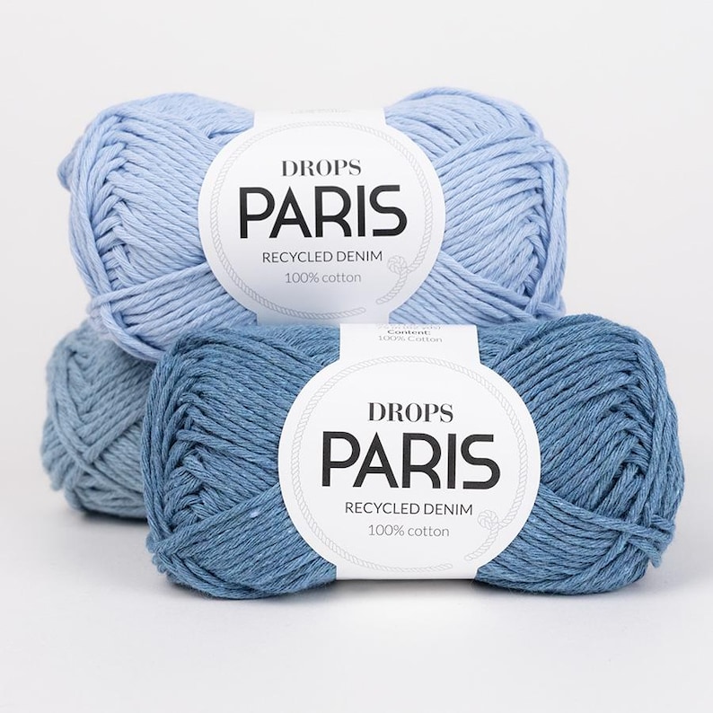K&ouml;nnte beinhalten: Zwei Kn&auml;uel Drops Paris Recycling-Denim-Garn in Blaut&ouml;nen. Die Etiketten zeigen "DROPS PARIS RECYCLED DENIM 100% Baumwolle."