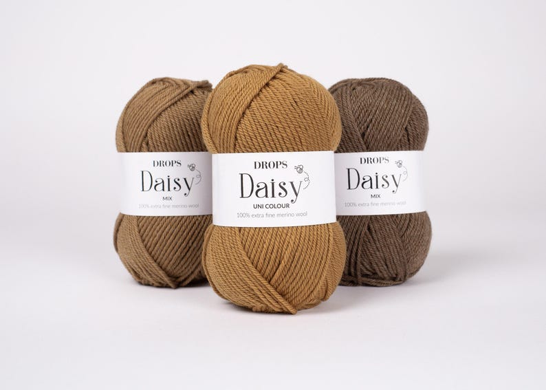 May include: Three skeins of yarn arranged in a row, featuring shades of light brown, medium golden brown, and dark brown. Each skein has a white paper label with the text DROPS Daisy, indicating 100% extra fine merino wool composition.