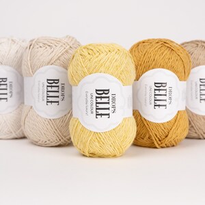May include: Five skeins of Drops Belle yarn in various colors, including cream, beige, yellow, and mustard. Each skein has a label with the text "BELLE" and "UNI COLOUR Everyday luxury!" against a white background. The yarn is arranged on a white surface.