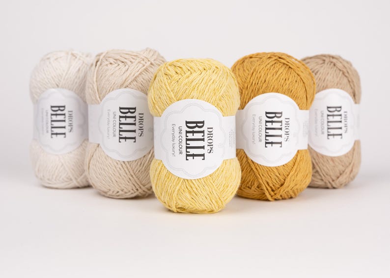 May include: Five skeins of Drops Belle yarn in various colors, including cream, beige, yellow, and mustard. Each skein has a label with the text "BELLE" and "UNI COLOUR Everyday luxury!" against a white background. The yarn is arranged on a white surface.