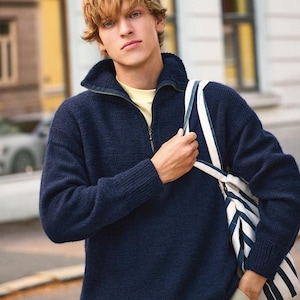May include: A man wearing a navy blue sweater with a zip and a white and blue striped tote bag.