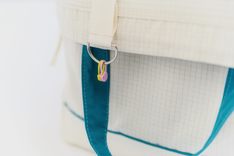 Puede incluir: Primer plano de un bolso tote blanco con detalles turquesa y un anillo de metal plateado. El bolso tiene un patr&oacute;n de cuadr&iacute;cula y un ribete turquesa. Al anillo est&aacute;n unidos dos peque&ntilde;os amuletos de pl&aacute;stico de colores amarillo y rosa.