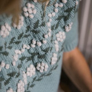 May include: A light blue knitted sweater with a floral pattern and white pom-poms. The sweater has short sleeves and a round neckline.