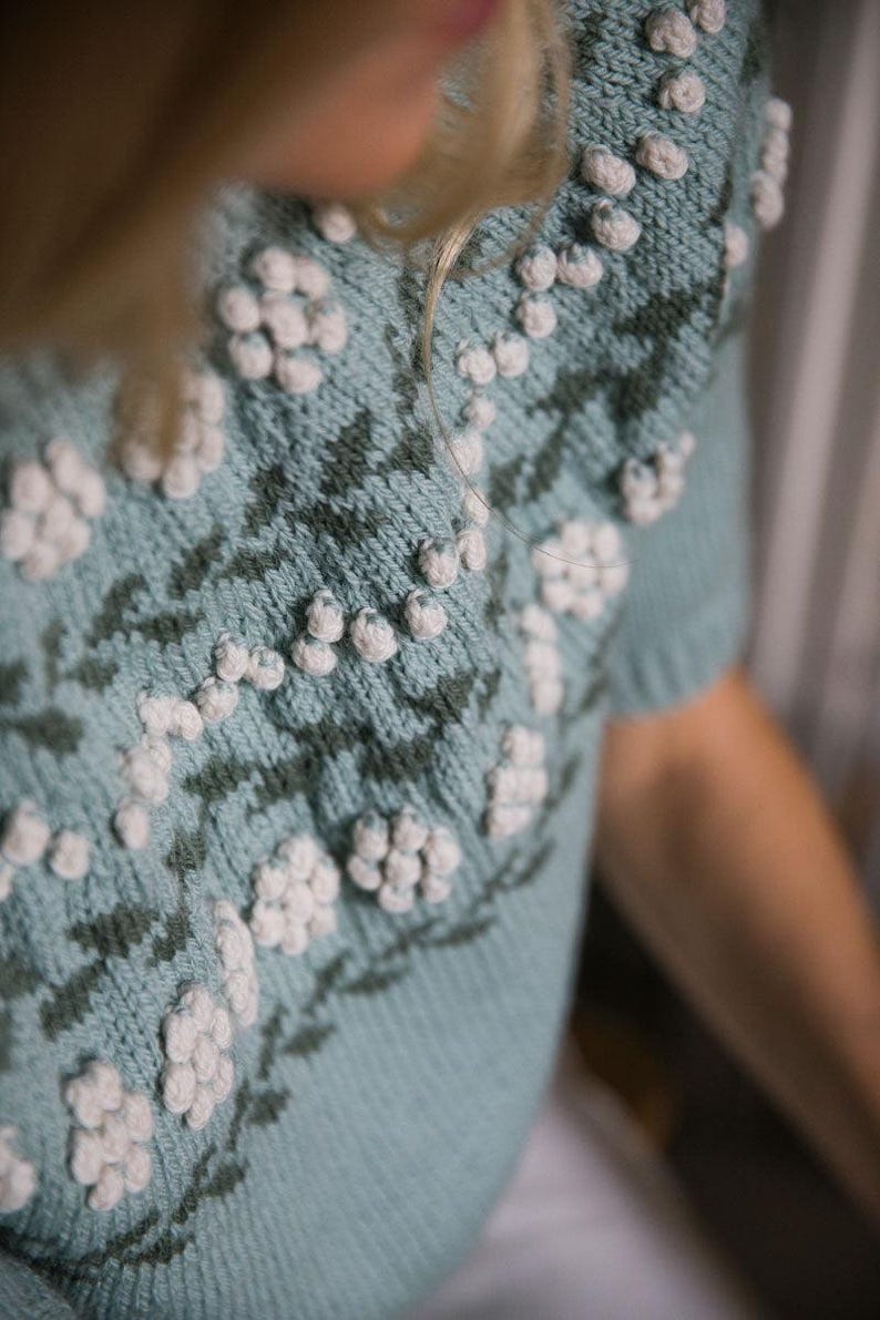 May include: A light blue knitted sweater with a floral pattern and white pom-poms. The sweater has short sleeves and a round neckline.