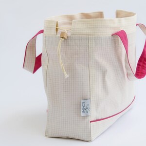 Puede incluir: Un bolso tote crema con un patr&oacute;n de cuadr&iacute;cula y detalles en rosa brillante. El bolso tiene asas y ribetes rosas, un cierre de cord&oacute;n y una peque&ntilde;a etiqueta con un logotipo de hoja.