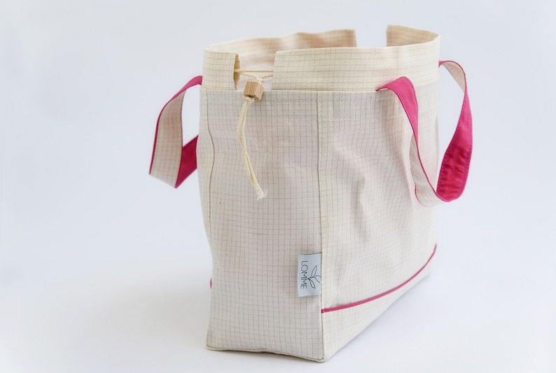 Puede incluir: Un bolso tote crema con un patr&oacute;n de cuadr&iacute;cula y detalles en rosa brillante. El bolso tiene asas y ribetes rosas, un cierre de cord&oacute;n y una peque&ntilde;a etiqueta con un logotipo de hoja.