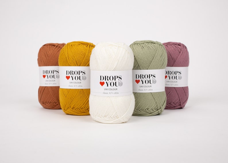 Puede incluir: Cinco ovillos de hilo Drops You Uni Colour en tonos &oacute;xido, amarillo mostaza, crema, verde salvia y malva. Cada ovillo tiene una etiqueta con el nombre de la marca y los detalles del producto. El hilo es algod&oacute;n cl&aacute;sico 8/4.