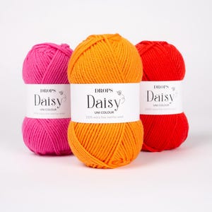 May include: Three skeins of yarn in bright pink, orange, and red. Each skein features a white paper label with the text DROPS Daisy UNI COLOUR 100% extra fine merino wool printed in black. The yarn is wound into a rounded, textured ball shape.