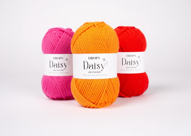 May include: Three skeins of yarn in bright pink, orange, and red. Each skein features a white paper label with the text DROPS Daisy UNI COLOUR 100% extra fine merino wool printed in black. The yarn is wound into a rounded, textured ball shape.