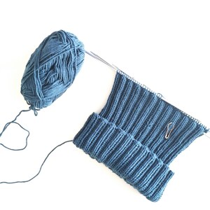Puede incluir: Un gorro de punto azul en progreso en agujas de tejer con un ovillo de lana azul.
