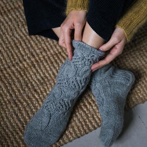 May include: A pair of gray knitted socks with a cable knit pattern. The socks are worn by a person sitting on a woven rug.