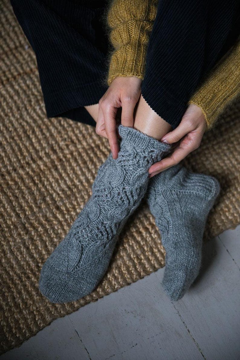May include: A pair of gray knitted socks with a cable knit pattern. The socks are worn by a person sitting on a woven rug.
